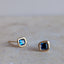 A pair of gold stud earrings with square-cut blue gemstones, resting on a light beige stone surface.