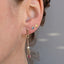 A close-up of a person's ear wearing a gold dangle earring with a small white pearl bead and a dangling pink gemstone, alongside two smaller gold stud earrings.