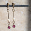 A pair of gold dangle earrings with white pearl beads and oval pink gemstone drops, hanging from a dark metal bar against a light beige stone background.