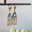 A pair of ornate gold-tone dangle earrings with oval iridescent blue opal stones, hanging from a black display bar against a light gray wall with green leaves at the bottom.