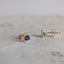 A pair of gold stud earrings with square-cut blue gemstones, one shown from the front and the other from the side/back, resting on a light beige stone surface.
