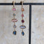 A pair of gold dangle earrings with multiple colorful gemstone accents, hanging from a black display stand against a beige stone-textured background.