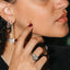Close-up of a person wearing silver jewelry, including a long dangling earring with a butterfly motif, a silver ring with a rectangular stone, and a delicate chain necklace, against a dark background.
