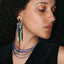 A close-up of a person wearing a long, multi-colored dangling earring with rectangular and cylindrical elements in metallic and pastel tones, shown against dark curly hair and skin.