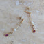 A pair of gold drop earrings with a small white pearl, a dangling oval red gemstone, and a round red gemstone at the end, displayed on a beige marble surface.