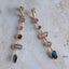 A pair of gold drop earrings with multiple colorful gemstones, displayed on a light beige stone surface.