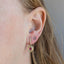 A close-up of a person's ear wearing a gold hoop earring with a spiral design and a small pink gemstone, alongside a smaller stud earring.