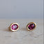 A pair of gold stud earrings with oval-cut pink gemstones, resting on a light beige stone surface against a soft gray background.