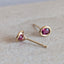 A pair of gold stud earrings with small oval settings and pink gemstones, resting on a textured beige stone surface.