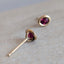A pair of gold stud earrings with oval deep pinkish-purple gemstones, resting on a textured light gray stone surface.