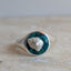 A silver signet ring with a round bezel containing a dark teal-green stone and a small white inlay, resting on a light beige stone surface.