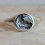 A silver signet ring with a textured, organic engraved face, resting on a light beige stone surface against a neutral gray background.