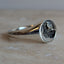 A silver ring with a textured, hammered oval face resting on a light beige stone surface against a neutral gray background.