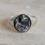 A silver signet ring with a dark, textured oval face featuring a bird design, resting on a light beige stone surface against a neutral gray background.