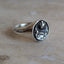 A silver signet ring with an engraved eagle emblem, resting on a light beige stone surface against a neutral gray background.