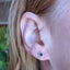 A close-up of a person's ear wearing a small square stud earring with a blue center and a gold border.