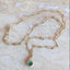 A gold chain necklace with a textured gold pendant holding a round green emerald, displayed on a light beige stone surface.