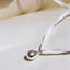 A silver teardrop pendant necklace with a white ribbon, resting on a warm beige surface under bright, direct sunlight.
