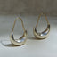 A pair of silver crescent-shaped hoop earrings with gold ear wires, displayed on a neutral beige surface with soft shadows.