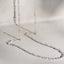 A delicate silver chain necklace draped on a light gray surface, showing the fine links and reflective metal against a soft gray background.