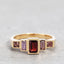 A gold ring featuring a central rectangular red gemstone flanked by two lavender baguette-cut stones and smaller square pinkish-purple stones on each side, set against a neutral gray background.