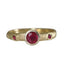 A gold ring featuring a central round-cut deep red gemstone in a bezel setting, with two smaller square-cut red gemstones on either side of the band, shown against a plain white background.